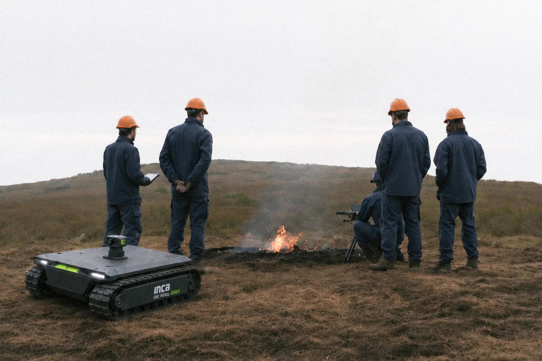 Training and research team in hard hats observing a controlled field fire, with an INCA Fire Patrol rover in the foreground
