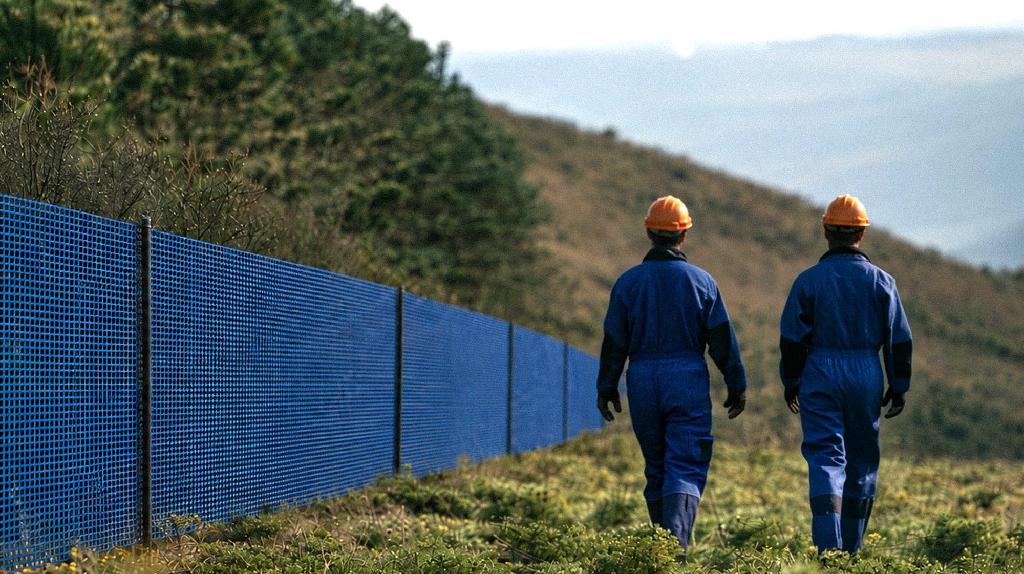 Two workers in blue coveralls and orange helmets walking beside an installed INCA WFS net® barrier on a hillside, with smoke rising in the distance