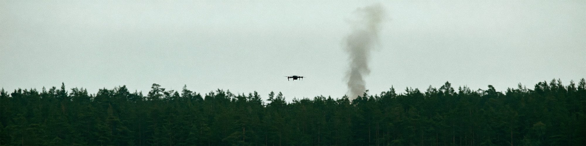 Forested ridge under overcast sky with a distant smoke column rising and a drone hovering above the treeline
