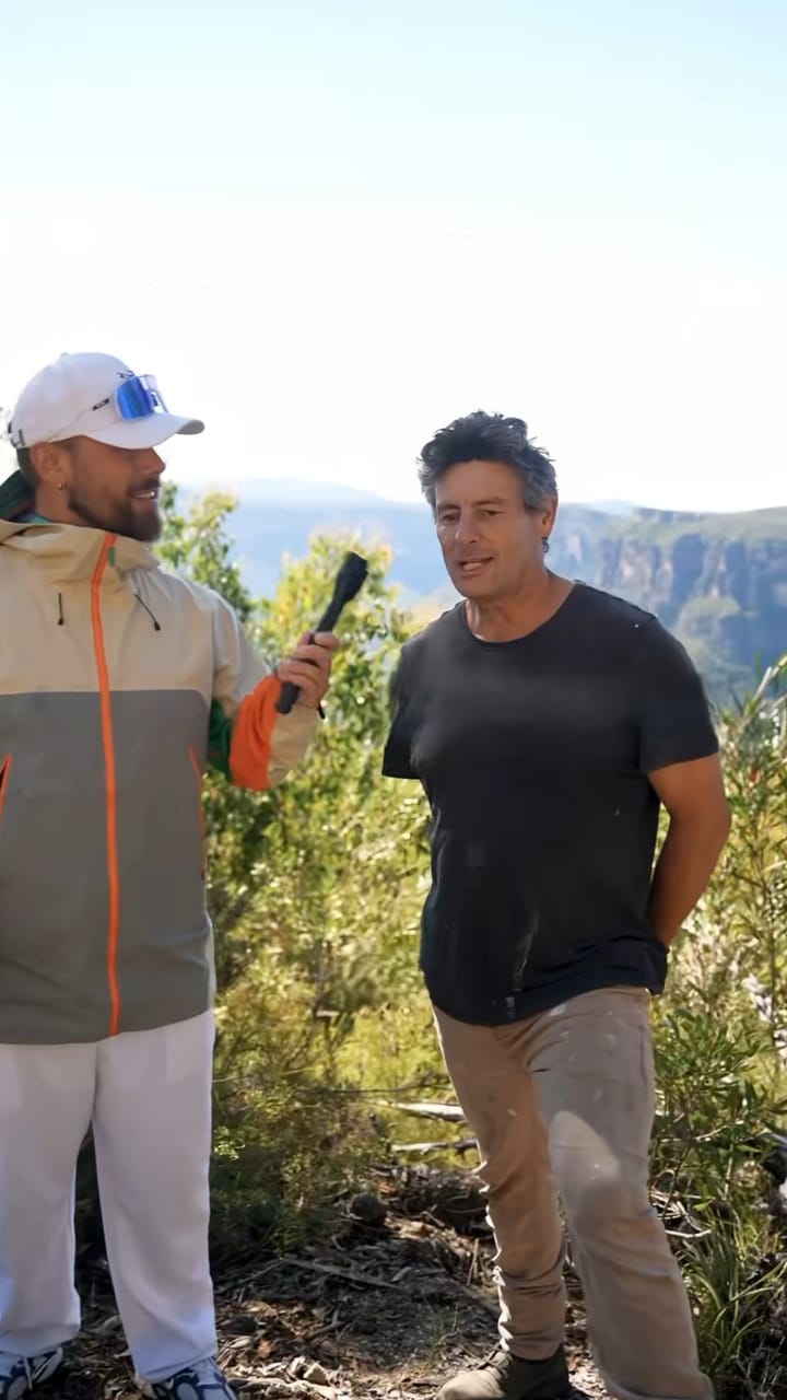 Presenter interviewing a man outdoors with hills behind