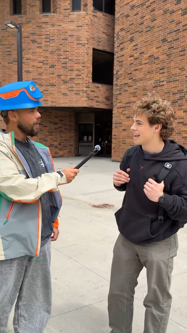 Presenter in a blue INCA helmet interviewing a young man outside a brick building
