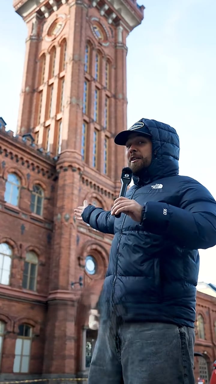Presenter in a dark jacket in front of the red-brick tower of Erottaja fire station in Helsinki