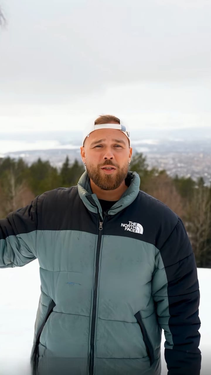 Presenter in a winter jacket on a hilltop with forest and a city in the distance